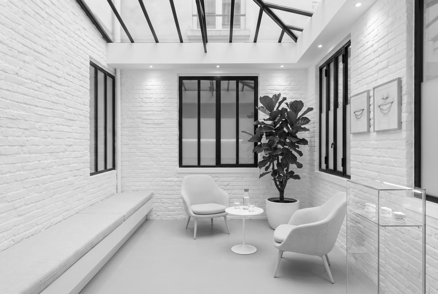 La salle d'attente sous la verrière : un banc et des fauteuils clairs, une table basse, un grand ficus et des dessins de sourires aux murs